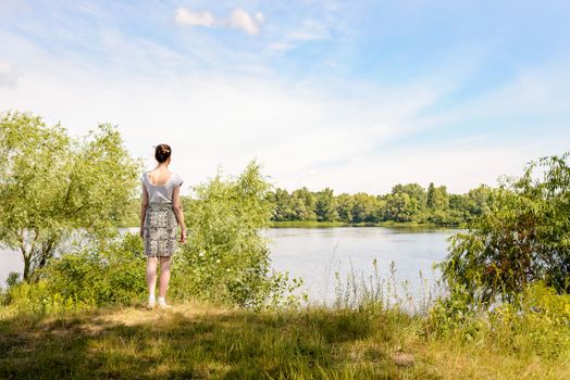 A woman standing up close to the Dnieper river in Kiev, Ukraine, observes in the distance from a raised point of view, under a soft cloudy summer sky