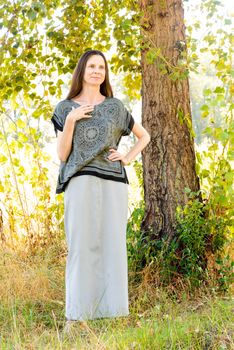 An elegant mature woman with a long dress in the shadow of a nice poplar tree in the park during a warm summer day