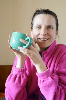 A smiling mature woman wearing a pink pull-over and drinking coffee in a green cup at morning for breakfast