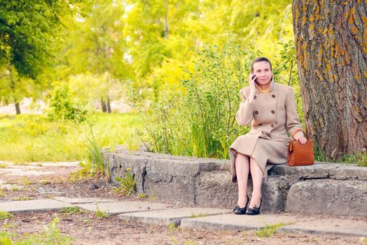 Elegant businesswoman sitting on a stone wall during a sunny spring day, and phoning to a customer