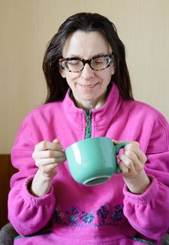 A smiling mature woman with glasses wearing a pink pull-over and drinking coffee in a green cup at morning for breakfast