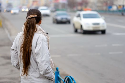 A woman with a ponytail stands, waiting or watching, close to the street with cars in the city