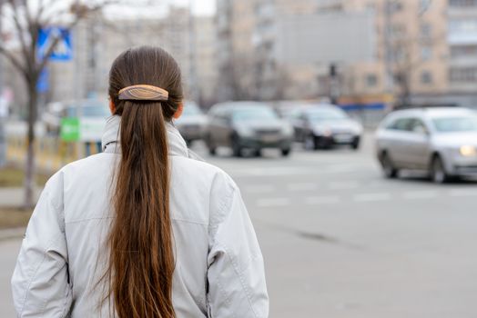 A woman with a ponytail stands, waiting or watching, close to the street with cars in the city