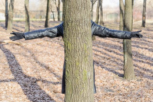 A woman wearing a black leather coat and gloves is hidden behind an oak tree in the park and spread her arms like a cross.