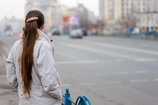 A woman with a ponytail stands, waiting or watching, close to the street with cars in the city
