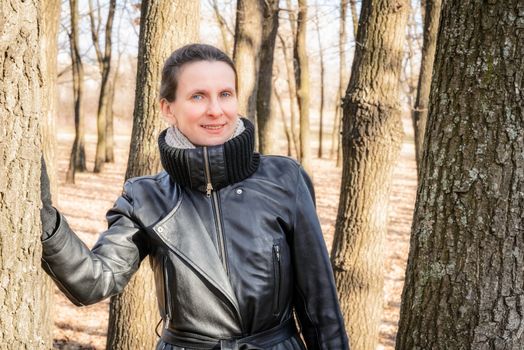 An adult woman wearing a black leather coat is standing in the oak trees forest
