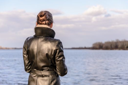 A woman with a chignon and a black leather coat is watching the landscape close to the lake or the river during a sunny day with big white clouds