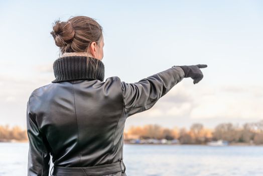 An adult with a chignon and wearing a black leather coat stays close to the river and point her finger to indicate something interesting on the opposite bank