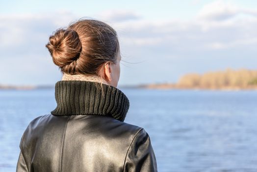 A woman with a chignon and a black leather coat is watching the landscape close to the lake or the river during a sunny day with big white clouds