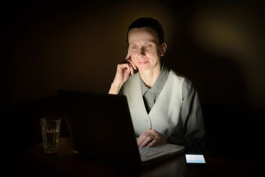 A business woman is using a computer in a dark room. Her face is illuminated by the light from the lcd monitor.