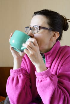 A mature woman with glasses wearing a pink pull-over and drinking coffee in a green cup at morning for breakfast