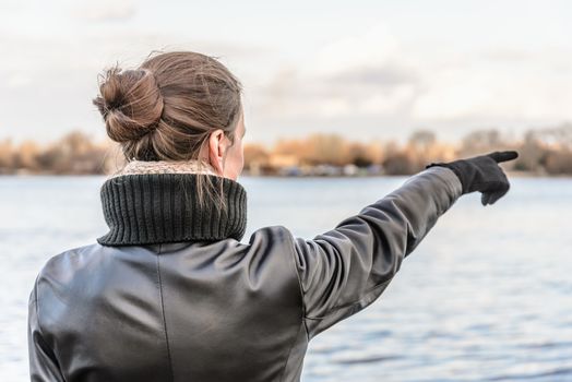 An adult with a chignon and wearing a black leather coat stays close to the river and point her finger to indicate something interesting on the opposite bank