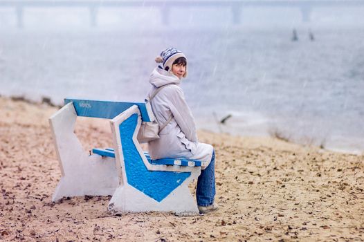 A woman sitting on a bench close to the river, in autumn under the rain