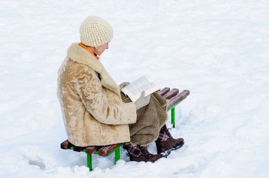 Woman sitting on a bench and reading a book in winter. Snow.