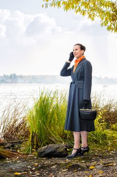 Elegant senior business woman with a mobile phone, a bag and an orange scarf, walking along the river, under the trees in autumn