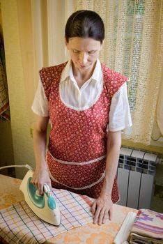 An adult woman, a housewife or a maid, wearing a red apron, is standing behind the ironing board. She irons some tea towels in the kitchen