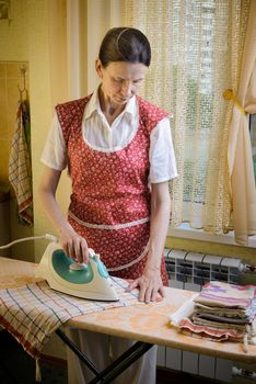 An adult woman, a housewife or a maid, wearing a red apron, is standing behind the ironing board. She irons some tea towels in the kitchen