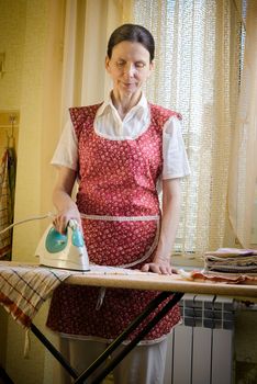 An adult woman, a housewife or a maid, wearing a red apron, is standing behind the ironing board. She irons some tea towels in the kitchen
