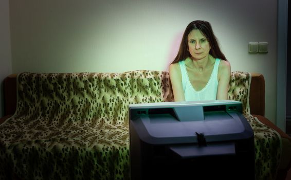 A middle-aged woman, alone, watches television, looking sad, sitting on a couch. She watches a sad movie or listens to bad news that makes her a little unhappy and depressed