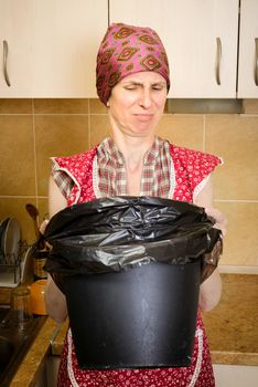 A woman, with a scarf on the head and a red apron, is looking inside a black trash can with a garbage bag, in the kitchen. She is very disturbed by the bad smell