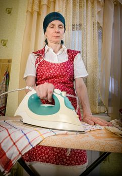 An adult woman, a housewife or a maid, with a green scarf on the head and wearing a red apron, is standing behind the ironing board. She irons some tea towels in the kitchen