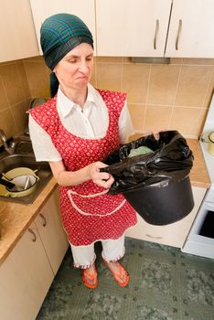 A woman, with a scarf on the head and a red apron, is looking inside a black trash can with a garbage bag, in the kitchen. She is very disturbed by the bad smell
