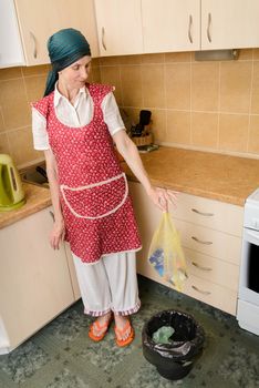 A woman, with a scarf on the head and a red apron, is looking inside a black trash, in the kitchen, holding a garbage bag in the hand. She is very disturbed by the bad smell