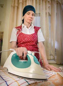 An adult woman, a housewife or a maid, with a green scarf on the head and wearing a red apron, is standing behind the ironing board. She irons some tea towels in the kitchen