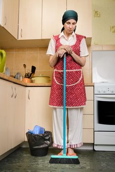 A depressed  adult woman, a housewife or a maid, wearing a red apron and a green scarf on her head is resting after she has swept the kitchen with a broom