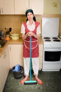 A depressed  adult woman, a housewife or a maid, wearing a red apron and a green scarf on her head is resting after she has swept the kitchen with a broom