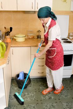 An  adult woman, a housewife or a maid, wearing a red apron and a green scarf on her head is sweeping the kitchen with a broom