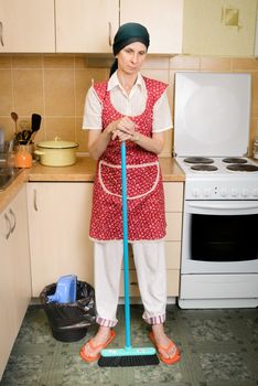 A depressed  adult woman, a housewife or a maid, wearing a red apron and a green scarf on her head is resting after she has swept the kitchen with a broom