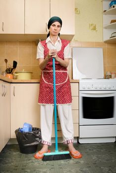 A depressed  adult woman, a housewife or a maid, wearing a red apron and a green scarf on her head is resting after she has swept the kitchen with a broom