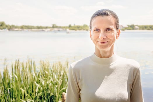 A warm portrait of a nice smiling senior woman wearing a white jumper close to the river in summer