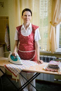 An adult woman, a housewife or a maid,  wearing a red apron, is standing behind the ironing board. She irons some tea towels in the kitchen