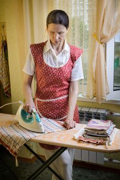 An adult woman, a housewife or a maid, wearing a red apron, is standing behind the ironing board. She irons some tea towels in the kitchen