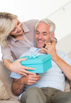 Portrait of a smiling woman surprising mature man with a gift on sofa at home