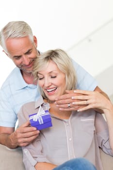 Portrait of a smiling mature man surprising woman with a gift on sofa at home