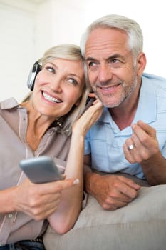 Cheerful mature man and woman with headphones and cellphone sitting on sofa at home