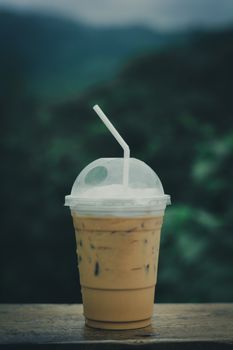 Ice coffee (espresso, cappuccino, latte) in plastic cup and white staw on wooden table with blurry green nature background. copy space for your text. Vacation and relaxing concept.