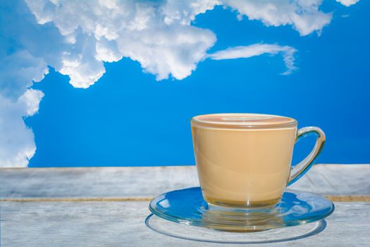 A cup of hot cappuccino or latte on wooden table and white clouds and blue sky background with copy space. Coffee times and relaxing concept.  