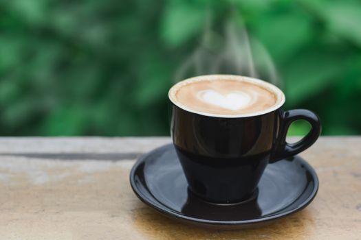 Hot coffee with smoke, heart shape with milk latte on top of black ceramic cup on wooden table in coffee shop and cafe with green background, copy space for your text. Times to relax with caffiene concept.