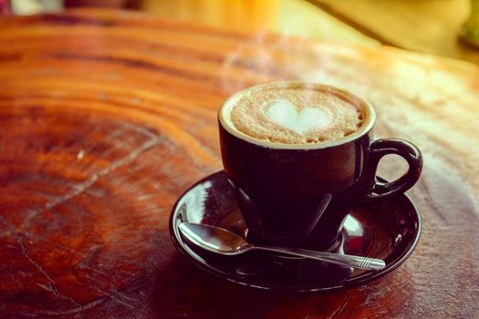 Hot coffee, heart shape with milk latte on top of black ceramic cup with metal spoon on wooden table in coffee shop and cafe in vintage tone, copy space for your text. Times to relax with caffiene concept.