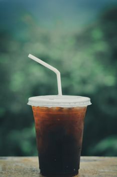 Ice coffee (espresso, cappuccino, latte) in plastic cup and white staw on wooden table with blurry green nature background. copy space for your text. Vacation and relaxing concept.