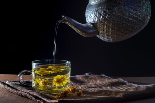 Pour water to a cup of Chrysanthemum tea with dry flower on brown table cloth on black background. Healthy beverage for drink. Herbs and medical concept.