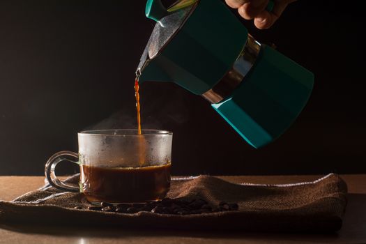 Pour hot of black coffee from green moka pot to clear coffee cup with smoke and coffee beans on brown table cloth and wooden table. Benefit of coffee concept.