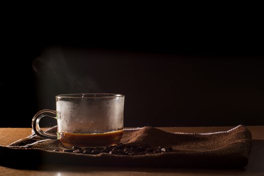 Cup of hot coffee with smoke and group of coffee beans on brown table cloth with black background with copy space for your text. Benefits of coffee concept.