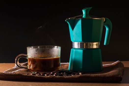 Close up hot of black coffee and green moka pot and coffee beans on brown table cloth and wooden table. Benefit of coffee concept. 