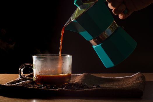 Pour hot of black coffee from green moka pot to clear coffee cup with smoke and coffee beans on brown table cloth and wooden table. Benefit of coffee concept.