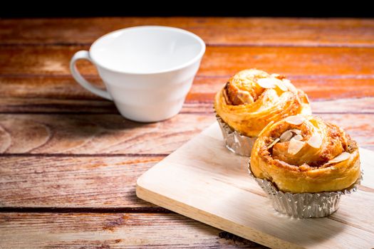 Delicious homemade almond danish bread in foil cup and white cup of coffee on a wooden chopping board and table in the home kitchen on black background.